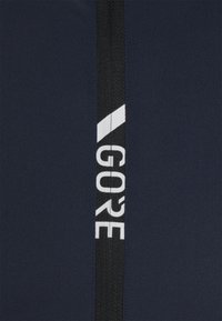 Marinblått tyg med ett strukturerat svart elastiskt band som har vit "GORE"-text och ett diagonalt vitt streck.