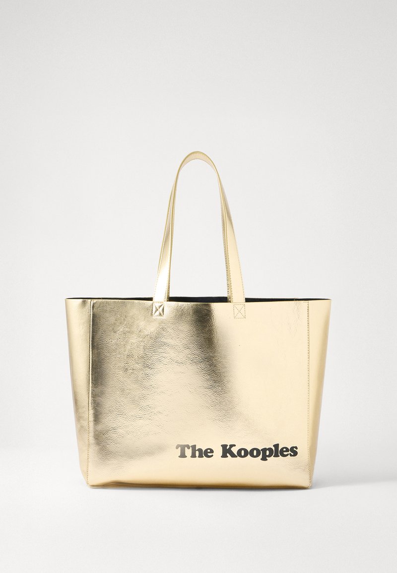 Metāliska zelta iepirkumu soma ar melnu apdari. Gluda tekstūra, strukturēts dizains un garas rokturi. Apakšā ir uzdrukāts "The Kooples".