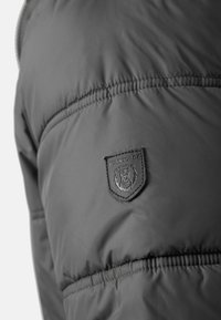 Section de veste matelassée gris foncé avec des coutures surpiquées et un petit patch en caoutchouc arborant un logo d'ours et le mot « JEANSCODE ».