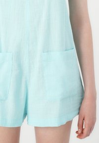 Jumpsuit bleu clair en tissu texturé, doté de deux poches plaquées à l'avant, avec une coupe décontractée et des jambes courtes.