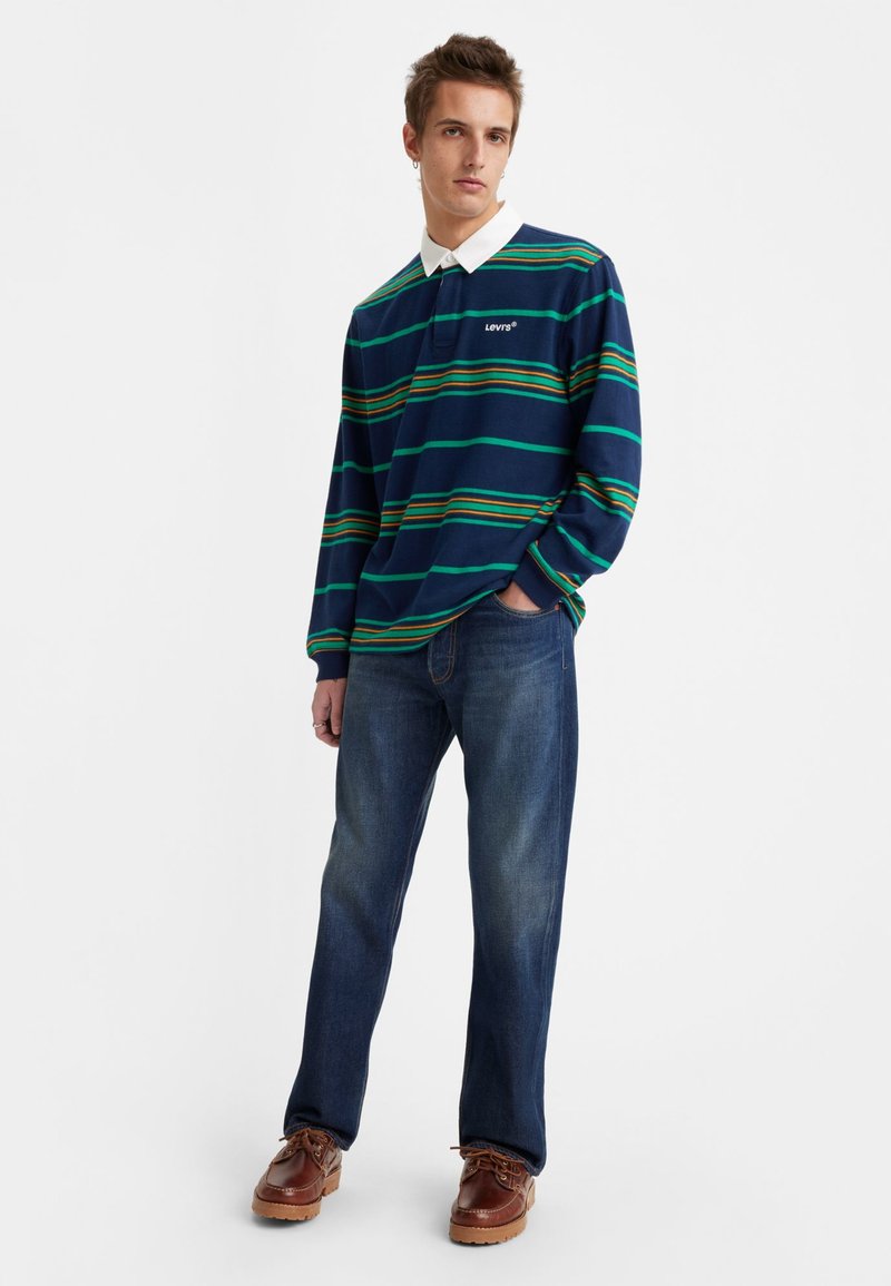 Navy-blau gestreiftes Langarm-Polo mit grünen und orangefarbenen Linien, weißem Kragen, kombiniert mit blauen Jeans und braunen Schnürschuhen.