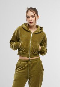 Sweat-shirt cropped à capuche en velours doux vert olive, avec poches kangourou et logo brodé. Associé à un pantalon assorti.
