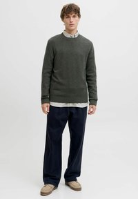 Grøn strikket sweater med rund hals, båret over en stribet skjorte, parret med løse marineblå bukser og lysebrune sko.