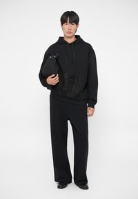 Melna kapuce ar priekšējo kabatu, kurai ir teksturēts raksts, apvienota ar brīva piegriezuma melnām biksēm. Modelis tur nelielu melnu somu.