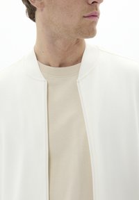 Chaqueta bomber blanca con cierre de cremallera y cuello acanalado, superpuesta sobre una camiseta de cuello redondo beige. Textura suave, diseño minimalista.