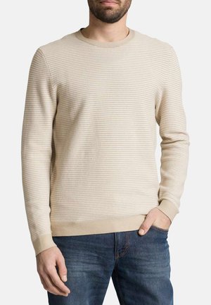 Mann trägt einen beigen, strukturierten Pullover mit langen Ärmeln und blaue Jeans, mit einer Hand in der Tasche, vor einfarbigem Hintergrund.