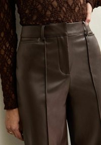 Close-up van bruine leren broeken met een hoge taille en riemlussen, gedragen met een gestructureerde donkerbruine langemouwen top.