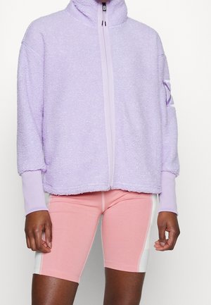 Personne portant une veste zippée en polaire violet clair avec des manches avec trous pour les pouces et un short de cyclisme rose et blanc à mi-cuisse.