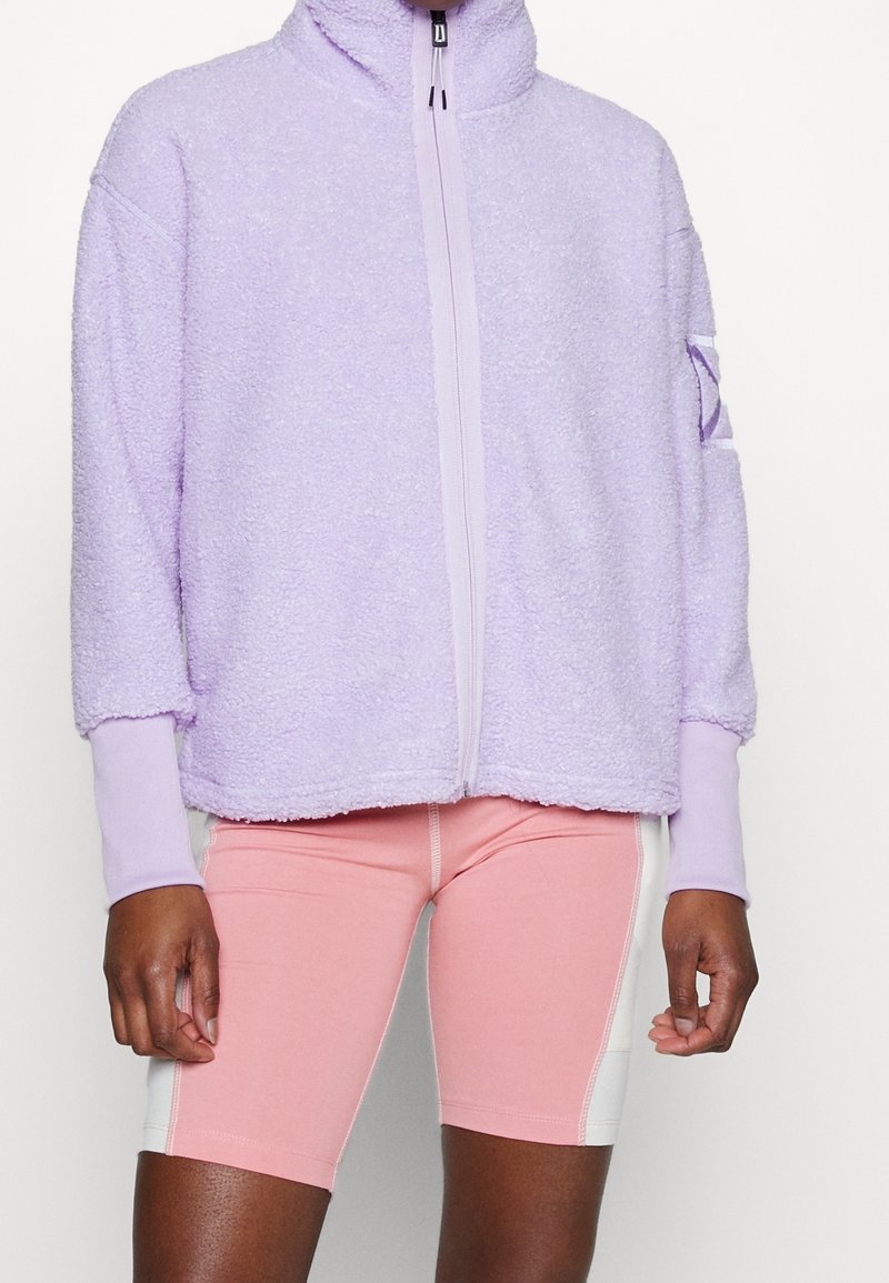 Personne portant une veste zippée en polaire violet clair avec des manches avec trous pour les pouces et un short de cyclisme rose et blanc à mi-cuisse.