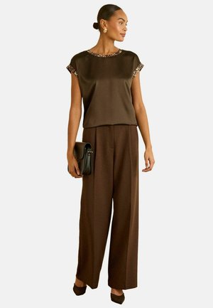 Braune Bluse mit Leopardenakzent am Ausschnitt, kombiniert mit hoch taillierter, weiter brauner Hose. Das Model hält eine schwarze Handtasche.