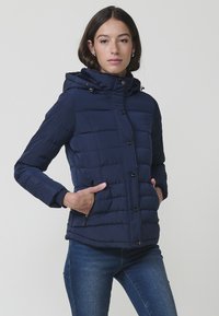 Tamno plava puffer jakna s kapuljačom, s kvadratnim uzorkom, prednjim gumbima i bočnim džepovima na zip. Glatki, vodoodbojni materijal.