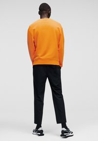 Oranssi huppari, mustat housut ja musta-valkoiset lenkkarit. Sileä kangas, normaali istuvuus, ribbaatut hihansuut ja helmassa, minimaalinen brändäys.