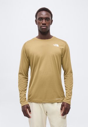Homme debout, face à l'avant, portant une chemise à manches longues beige avec le logo "The North Face" et un pantalon de couleur claire.