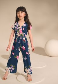 Navy cvetlični kombinezon s kratkimi rokavi, z rožami v roza in beli barvi, z elastičnim pasom in širokimi hlačnicami. Kombiniran z lahkimi roza sandali.