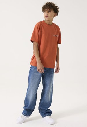Rostorange Baumwoll-T-Shirt mit kurzen Ärmeln und rundem Halsausschnitt, kombiniert mit locker sitzenden blauen Jeans und weißen Sneakers.