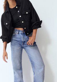 Zwarte spijkerjas met zilveren knopen, twee borstzakken, opgerolde mouwen, gecombineerd met lichtblauwe high-waisted jeans met rechte pijpen.