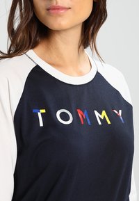 Dark navy sweatshirt with white raglan sleeves, featuring multicolored block letters spelling "TOMMY" on the front. Soft cotton blend fabric.