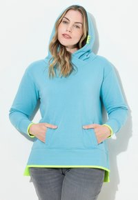 Hoodie azzurro chiaro con un grande cappuccio, tasche frontali e orlo posteriore più lungo. Presenta rifiniture verde neon sui polsini e sull'orlo inferiore. Texture morbida.