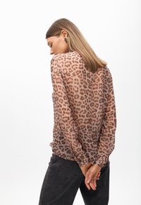 Blusa trasparente a maniche lunghe di un leggero rosa con stampa maculata marrone. Presenta polsini arricciati e una vestibilità ampia, abbinata a jeans scuri.