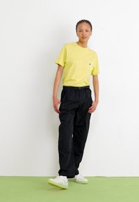 Gelbes kurzärmliges T-Shirt mit einer Brusttasche, kombiniert mit lockeren schwarzen Hosen. Die Schuhe sind weiße Sneakers mit grünen Sohlen.
