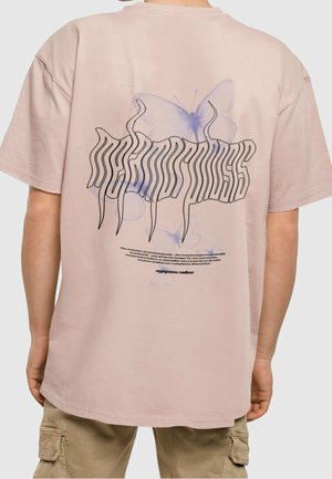 Osoba nosząca beżowy t-shirt z wieloma przezroczystymi fioletowymi nadrukami motyli oraz zdeformowanym czarnym tekstem na plecach.