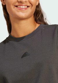 Camiseta de algodón gris oscuro con cuello redondo y puños acanalados, que presenta un logo de Adidas en negro bordado en el pecho. Textura suave.