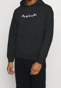 Sudadera negra con bolsillo frontal, capucha con cordón y gráfico "Light Addict" en púrpura y blanco. Fabricada en un tejido suave y liso con puños acanalados.