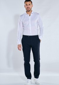 White button-up shirt with long sleeves, paired with navy slim-fit trousers and white sneakers, displayed on a neutral background.