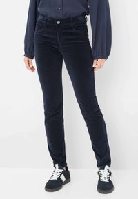 Pantalons en velours côtelé marine avec coupe slim, poches avant et fermeture à bouton. Associés à des chaussures de sport sombres avec des accents blancs.
