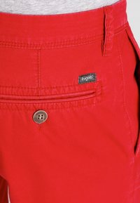 Punaiset shortsit, joissa on sileä pinta, varustettuna läppätaskulla ja pyöreällä metallinapilla. Brändilabel "bugatti" mustalla värillä taskussa.