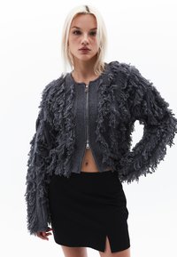 Grå fransig cardigan med ribbad textur, zip-stängning och lös passform. Designen har långa, fransiga ärmar och en kort längd.