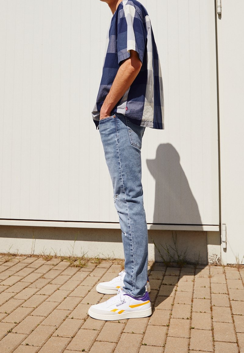 Homme portant une chemise à carreaux bleue et blanche, un jean bleu clair et des baskets blanches, debout sur une surface pavée avec les mains dans les poches.