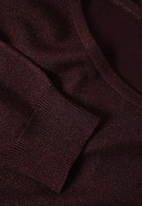 Gros plan sur un pull bordeaux, présentant une texture lisse, un col rond, et une poche plaquée avec un bord côtelé. Tissu doux avec un éclat subtil.