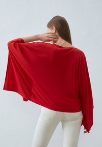 Rotes, fließendes Batwing-Oberteil aus glattem Stoff, mit einem weiten Ausschnitt und elegant über den Rücken fallend, kombiniert mit weißen, eng anliegenden Jeans.