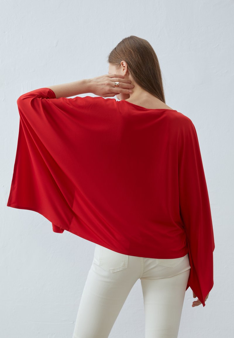 Top batwing rosso e fluente realizzato in tessuto morbido, caratterizzato da un ampio scollo e che si adagia elegantemente sulla schiena, abbinato a jeans bianchi aderenti.