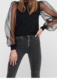 Haut noir avec manches transparentes, bouffantes et en tulle, associé à un jean gris foncé taille haute avec une patte de boutonnage à cinq boutons à l'avant.