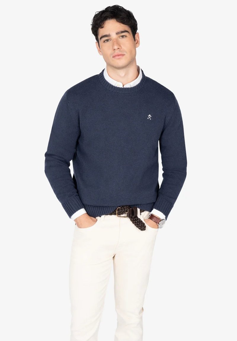 Jersey de color azul marino de punto con puños y dobladillo acanalados, con un pequeño logo en el pecho, combinado con pantalones beige claro y un cinturón trenzado.