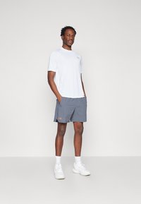 Weißes sportliches T-Shirt mit kleinem Logo, kombiniert mit grauen Shorts, die ein oranges Logo tragen. Das Model steht vor einem neutralen Hintergrund.