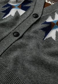 Grauer gestrickter Cardigan mit V-Ausschnitt, schwarzen Knöpfen und geometrischen Mustern in Blau, Weiß und Braun. Weiche Textur ist sichtbar.