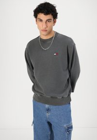 Felpa grigia con colletto e polsini a coste, caratterizzata da una piccola etichetta logo in rosso, bianco e blu. Abbinata a jeans cargo blu.
