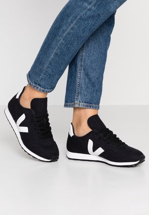 Une personne portant des baskets noir et blanc avec le logo "V", associées à un jean bleu, se tenant sur un sol blanc devant un fond uni.