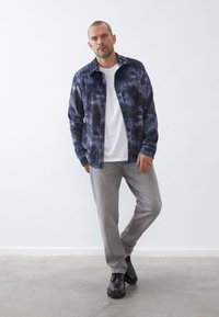 Camisa de pana de teñido anudado en color navy, camiseta blanca debajo, pantalones grises y zapatos negros; pose de pie con las manos en los bolsillos contra un fondo neutro.