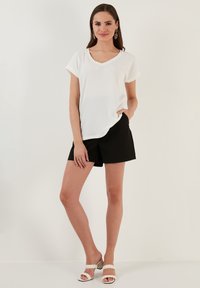 Top bianco a V con maniche corte, abbinato a pantaloni corti neri. Modello che indossa sandali bianchi con cinturini intrecciati, in piedi su uno sfondo chiaro.