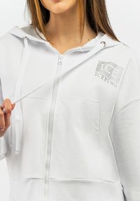 Felpa bianca con zip e tasca frontale, caratterizzata da un logo testurizzato in dettaglio scintillante. Il tessuto sembra morbido e leggero, con cappuccio.