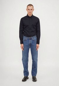 Zwarte knopen-overhemd gemaakt van gladde stof, gecombineerd met blauwe rechte jeans. Schoenen zijn van zwart leer met een zachte textuur.