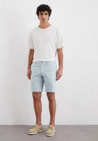 Shorts bleu clair avec une texture subtile, une fermeture à bouton et à zip, associés à un t-shirt blanc uni et des chaussures beiges à lacets.