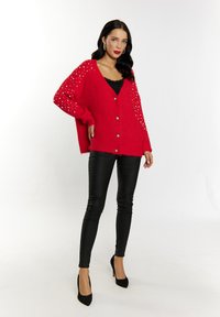 Cardigan rosso lavorato a maglia con scollo a V, chiusura a bottoni e decorazioni argento sulle maniche. Abbinato a pantaloni skinny neri e décolleté.