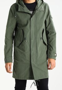 Parka verte avec capuche, en tissu léger, dotée de deux poches avant, de cordons de serrage réglables et de accents de boutons sur les poignets.