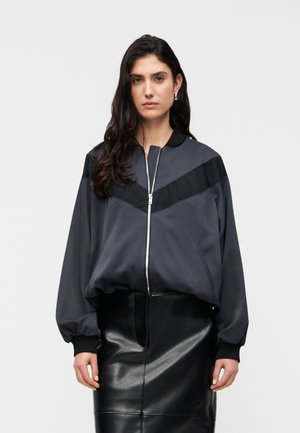 Dark gray zip-up jacket with a contrast black panel across the chest, paired with a shiny black leather skirt. Soft texture, relaxed fit.