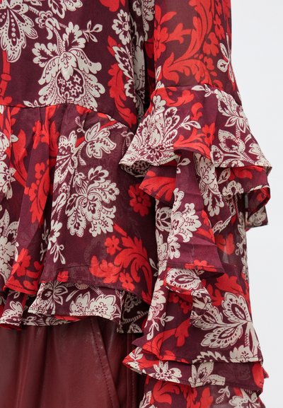 Pruunikarva lilleline pluus läbipaistvast kangast, millel on punased ja kreemjad mustrid. Volangidega varrukad kihilise tekstuuriga. Elegantne disain.